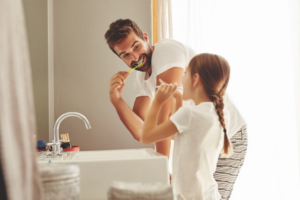 Father and daughter smiling at each other as they brush their teeth